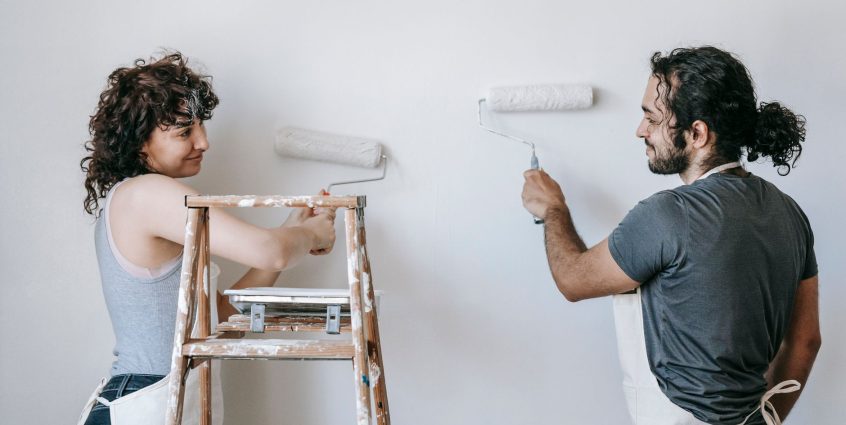Cheerful adult ethnic couple in aprons looking at each other while painting wall with rollers during renovation process in ho