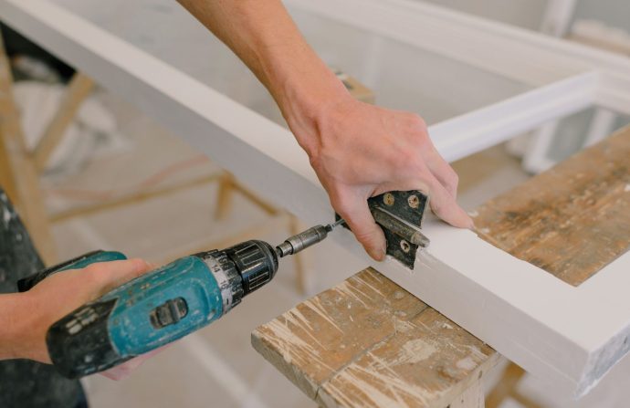 Unrecognizable professional male master using cordless screwdriver to install metal hinge on wooden window in light room duri