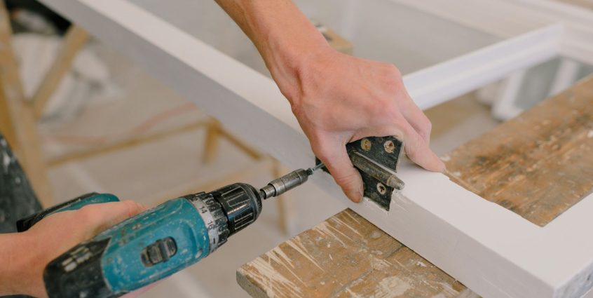 Unrecognizable professional male master using cordless screwdriver to install metal hinge on wooden window in light room duri