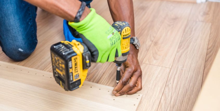 Close-up of handyman drilling wood indoors with green gloves and cordless drill. — Clover App Features Dating on Need Element
