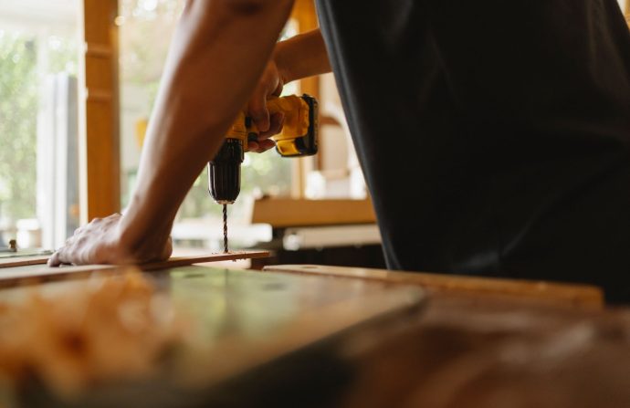 Crop anonymous male carpenter making hole on wooden plank with drill during work in studio on sunny day — Digital News and Ti