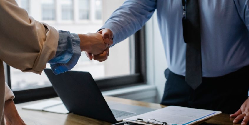 Crop unrecognizable coworkers in formal wear standing at table with laptop and documents while greeting each other before mee
