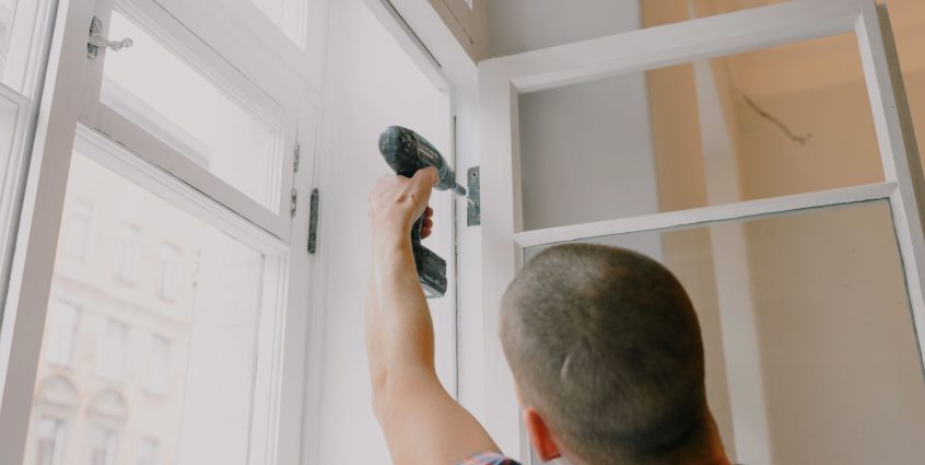 Back view of unrecognizable craftsman using cordless screwdriver and assembling window while screwing metal hinge during reno