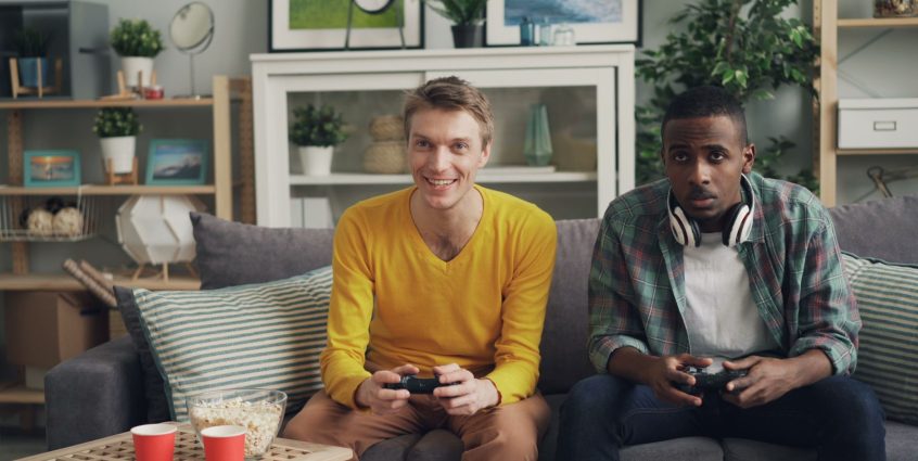 Two friends playing video games together on a sofa, one holding a controller and the other wearing headphones. — How Scam Sit