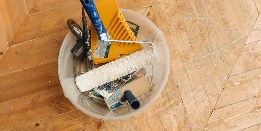 A collection of home renovation tools in a bucket on a wooden floor, ready for a painting project. — How to Buy Essays Online