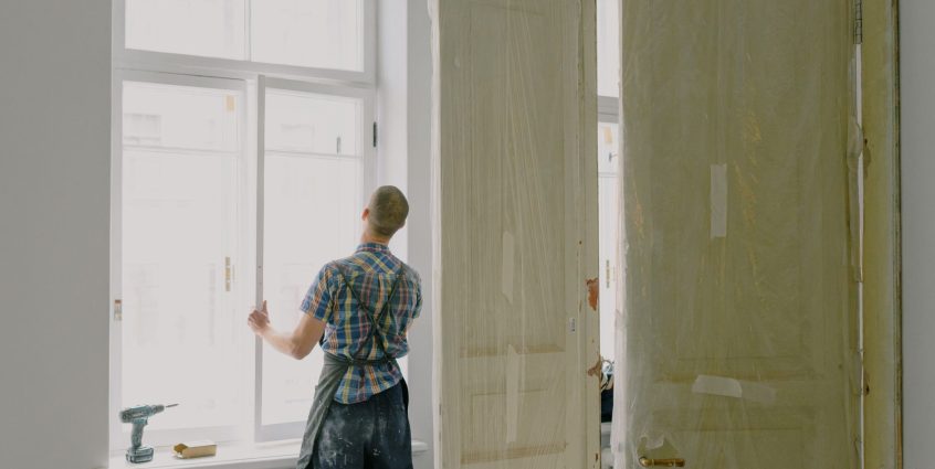 Back view of unrecognizable workman in apron mounting window near drilling machine and doors in flat — How to locate Energy f