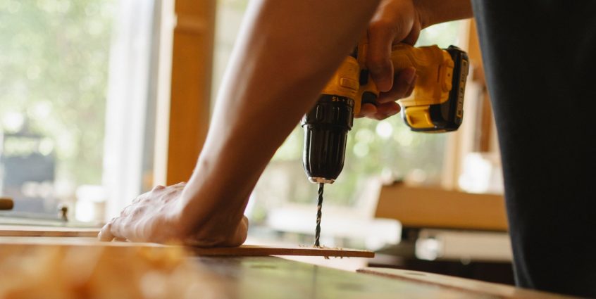 Crop anonymous male drilling timber plank with professional screwdriver on table in workshop — Research Paper Writer For Hire