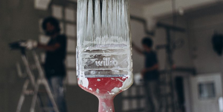 Detailed close-up of a paintbrush with men painting a wall in the blurred background, highlighting renovation work. — test