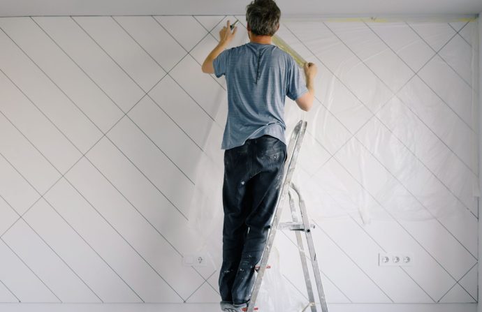 Full body back view of faceless male in shirt and dirty pants repainting wall while standing on ladder in bright empty room i
