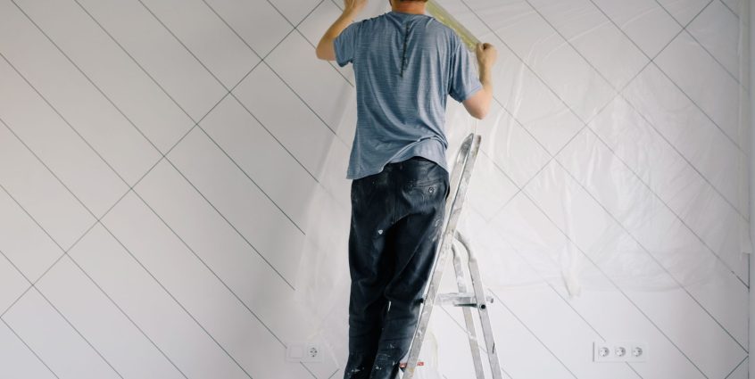 Full body back view of faceless male in shirt and dirty pants repainting wall while standing on ladder in bright empty room i